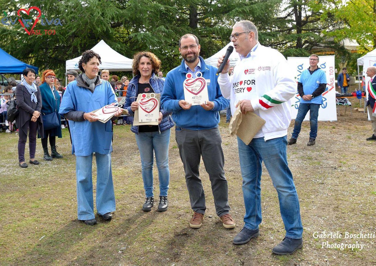 Premiazione all'aperto: quattro persone con targhe a forma di cuore, una parla al microfono